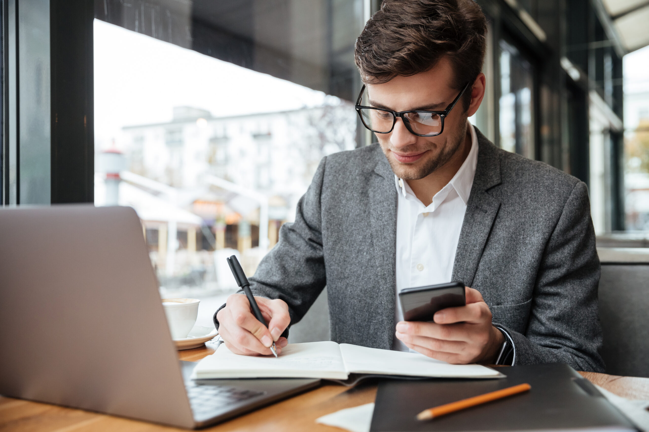 smiling-businessman-eyeglasses-sitting-by-table-cafe-with-laptop-computer-while-using-smartphone-writing-something Home